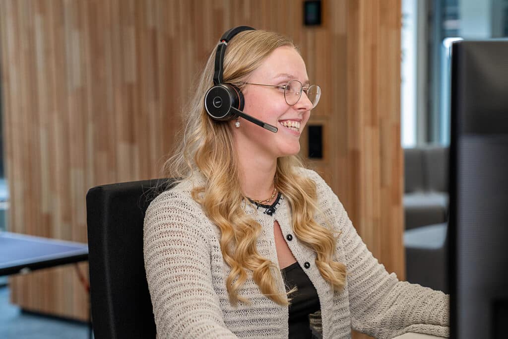 Eine junge blonde Frau sitzt mit Headset an einem PC und lacht.